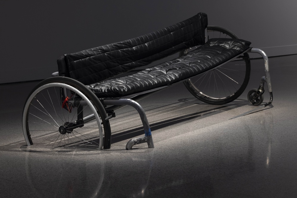 This image shows a bench with a sculptural shape that clearly resembles a mobility aid. The seat and backrest are padded in black, providing a comfortable, soft, quilted feel that stands out against the harder materials of the frame. The structure of the bench is made from metal tubing, giving it a modern, industrial look. On each side, there are large wheels similar to those found on wheelchairs. The bench is placed on a smooth, painted grey floor.