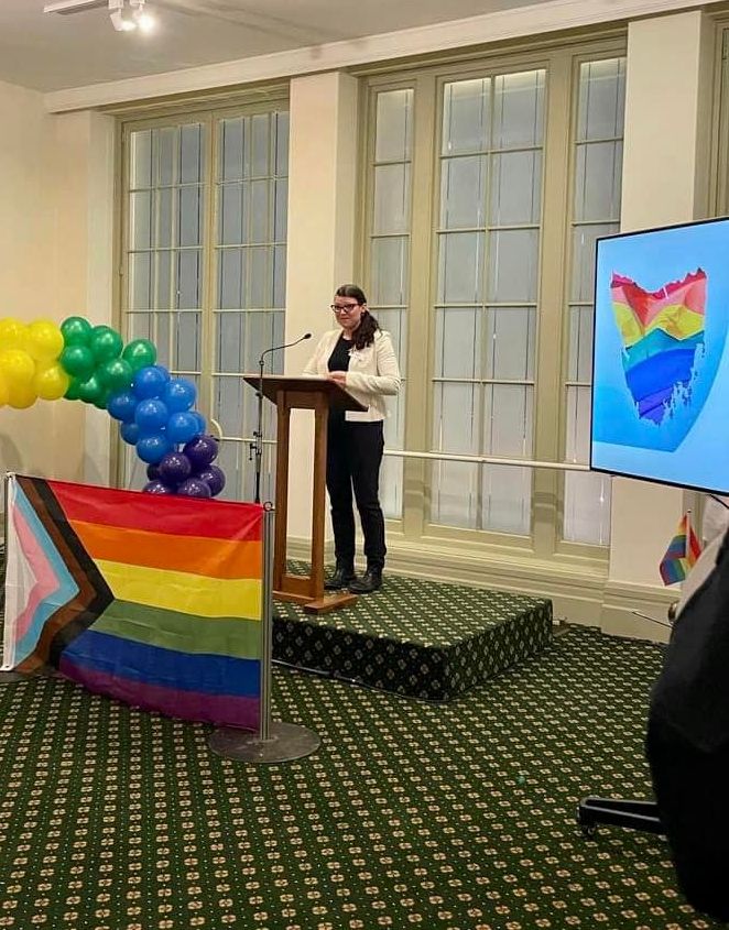 Angela is standing behind a lectern speaking. In the foreground is a pride flag. To Angela's left is a map of Tasmania projected on a screen. The map is filled with pride colours. To Angela's right are balloon in an arch shape in rainbow colours.  