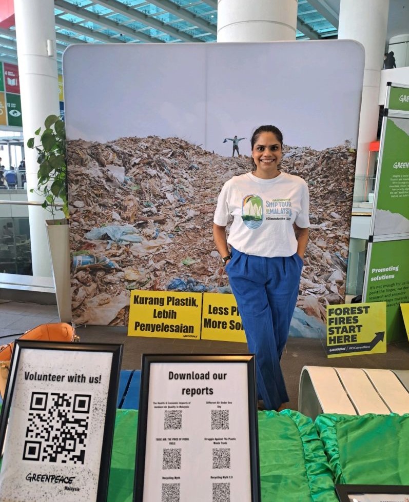 University of Tasmania alumna Hema Mahadevan in her role as a public engagement campaigner for Greenpeace Malaysia, standing with her hands in her pockets, smiling, wearing a Greenpeace t-shirt, surrounded by campaign posters.