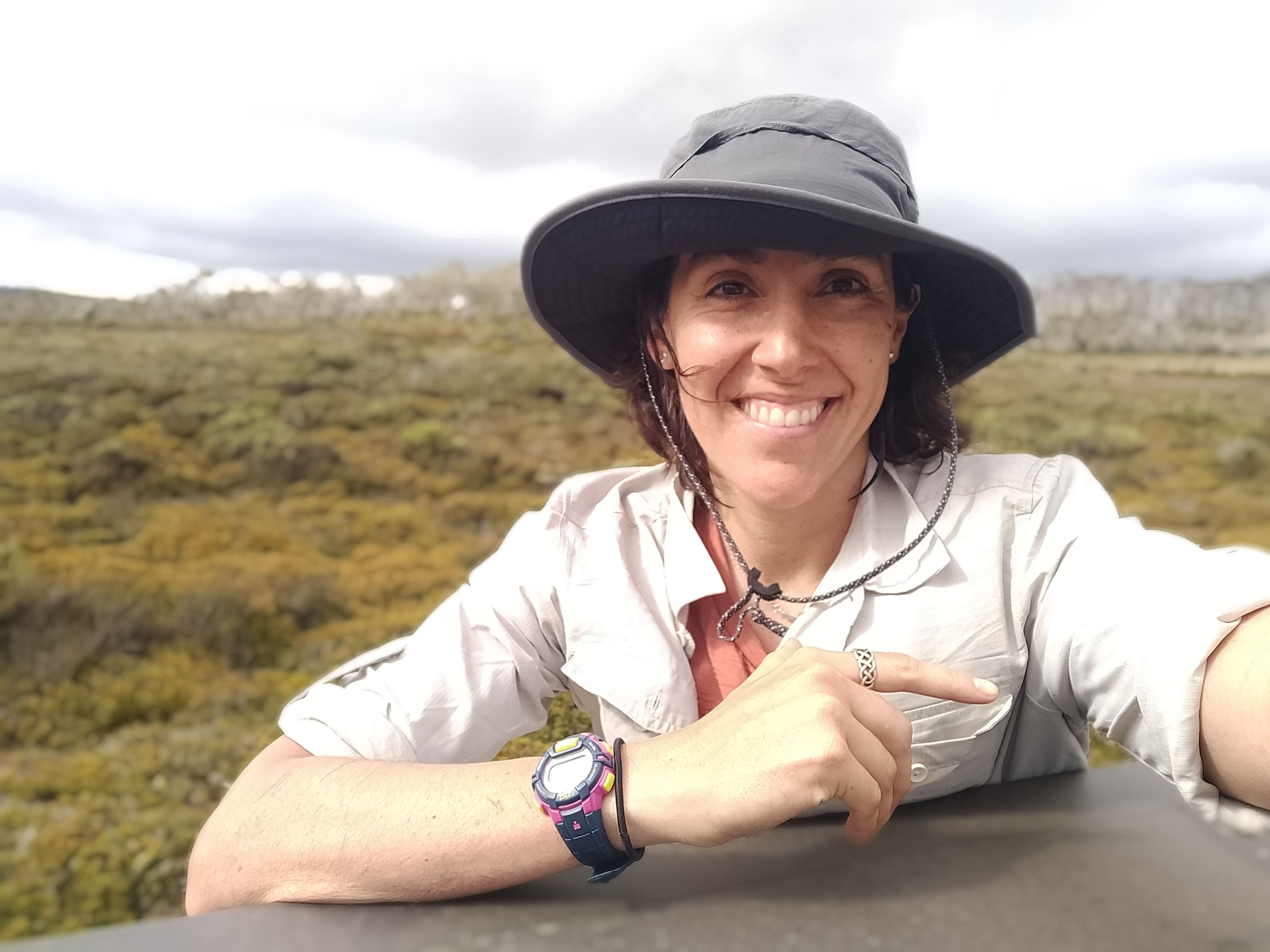 Laura N. Sotomayor in a wide-brimmed hat and light-colored clothing standing in a natural setting with hills and vegetation.