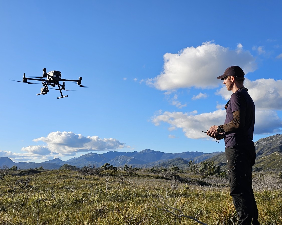 Arko operating a drone in a grassy field with mountains in the background under a partly cloudy sky.