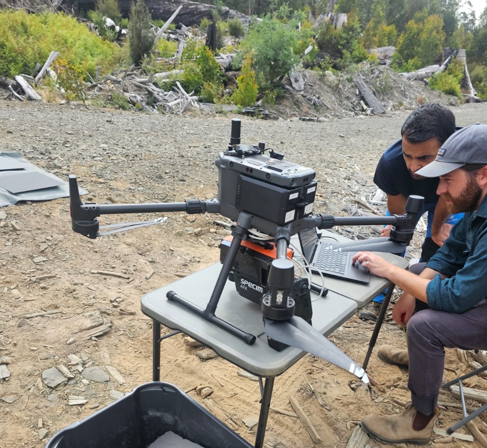 Two people work with a sensor-equipped drone on a table outdoors near rocky terrain and logs.