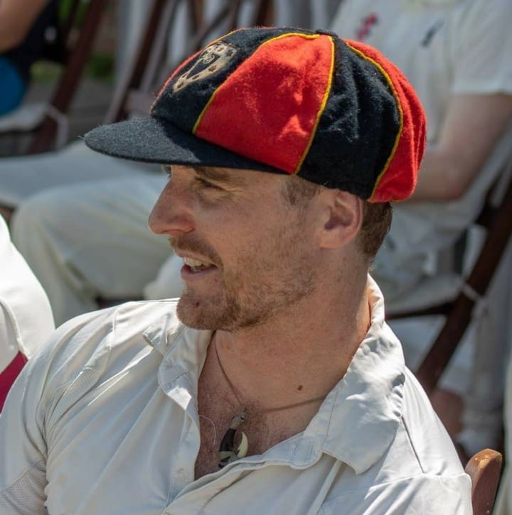 Robert wears cricket whites with an open-collared shirt and a red-and-black 