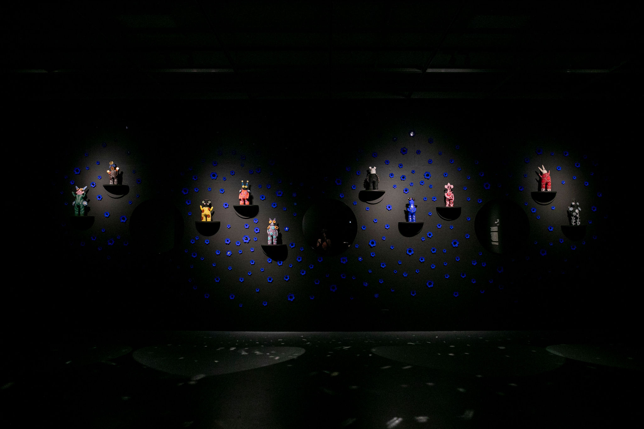 Image of a darkened gallery installation featuring a row of illuminated ceramic artworks by artist Vipoo Srivilasa. The works are mounted on a wall and surrounded by scattered patterns of small blue ceramic flowers. Three large circular mirrors are positioned among the artworks, creating reflective focal points within the display. Focused lighting highlights each ceramic piece, while the surrounding gallery remains in shadow.