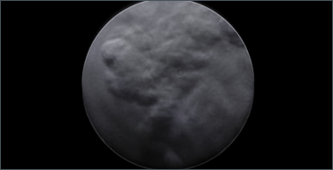 Against a completely black background, there is a large grey circle positioned in the centre of the image. The circle fills most of the frame and resembles a moon or a planet. Its surface is uneven and mottled, with soft swirls and patches of light and dark grey that look like clouds, smoke, or weather patterns moving across it. There are no sharp edges or distinct features, only subtle variations in tone and texture that create a sense of depth. The overall appearance is quiet, mysterious, and atmospheric.