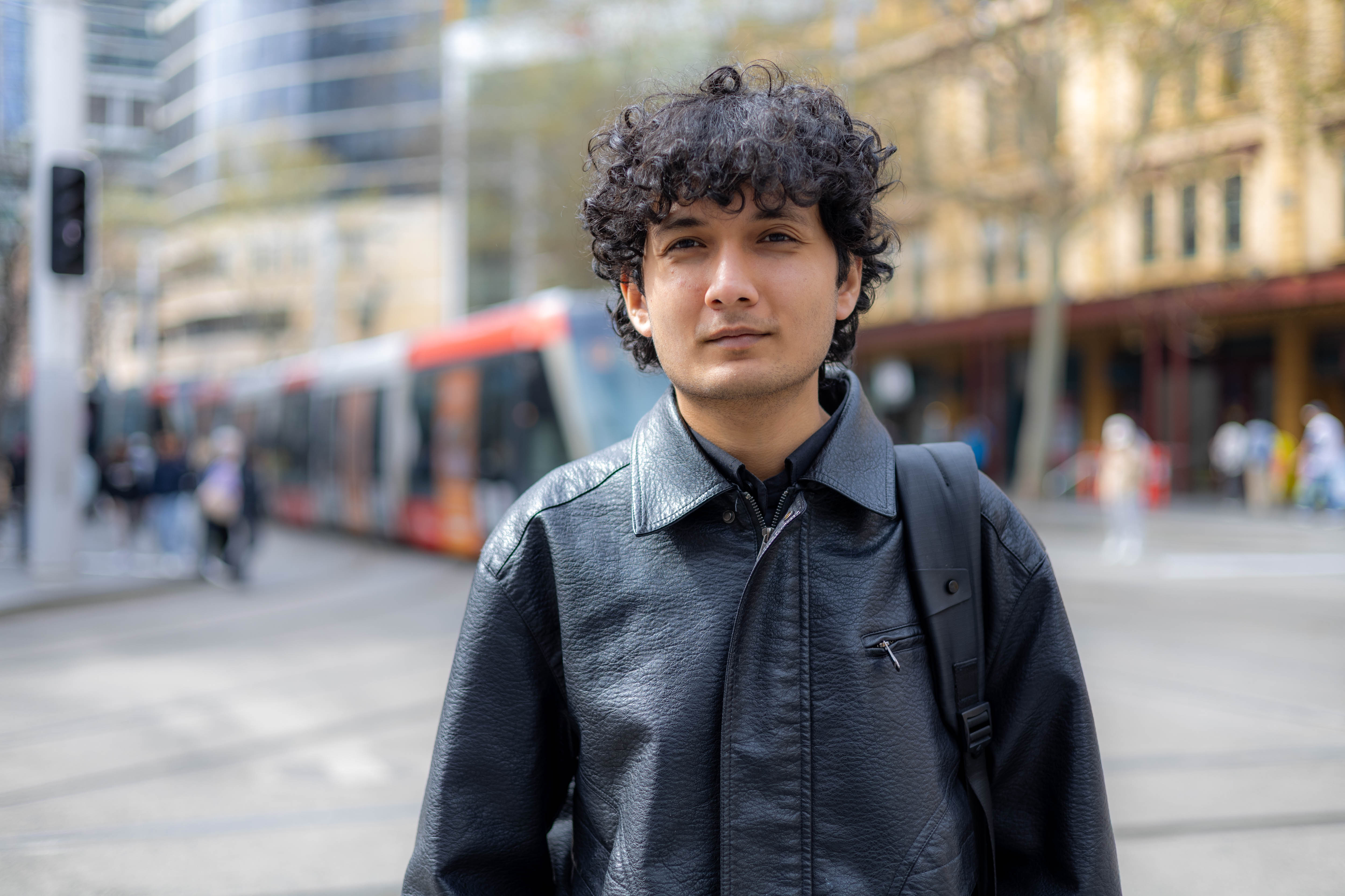 A young Nepalese man, with brown eyes and black curly hair, is standing in a Sydney street, looking at the camera with a slight smile. He is wearing a black leather jacket, the strap of a black backpack over one shoulder, and a red-and-white tram is passing in the background.