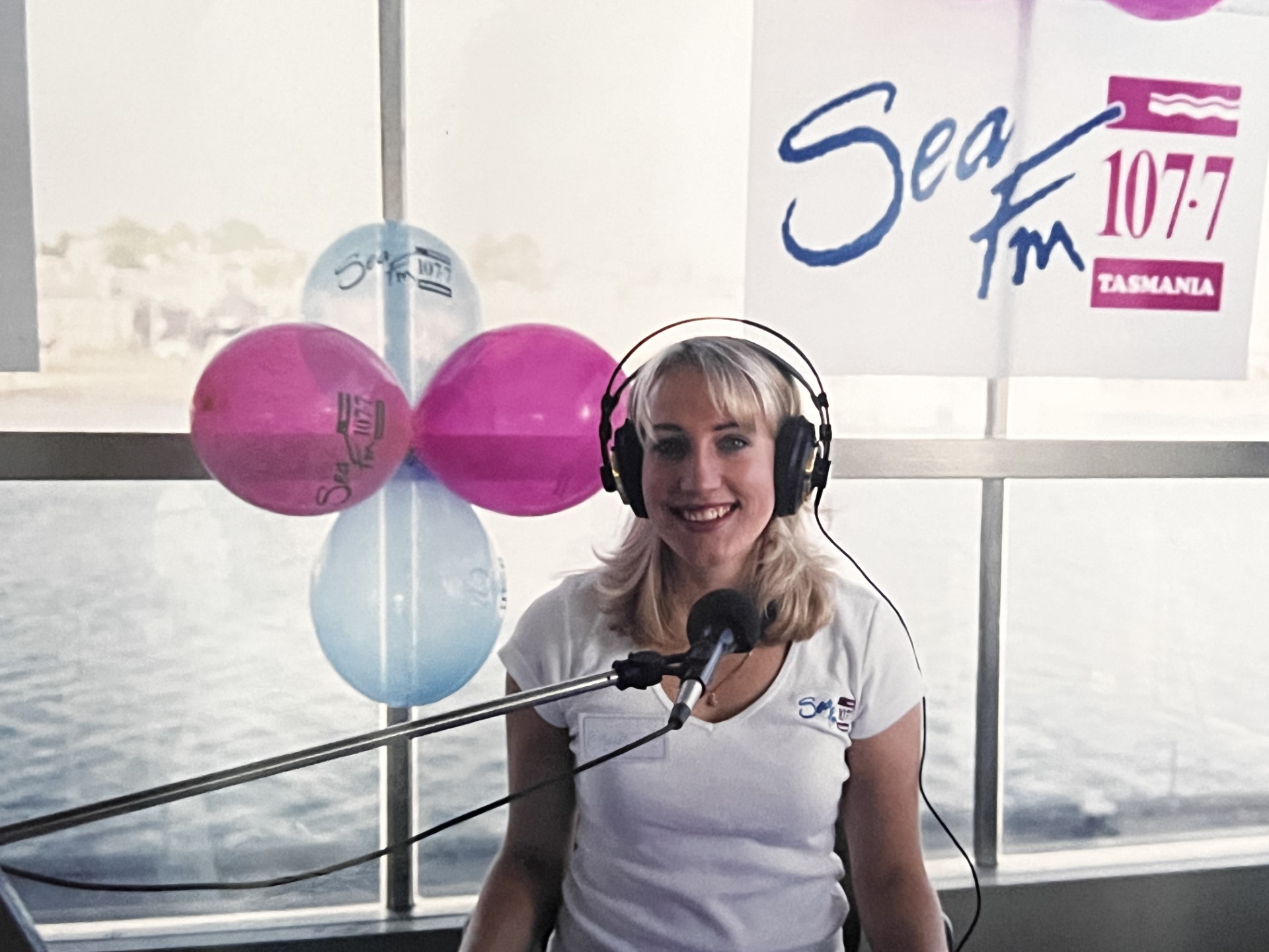 Emily Jade O'Keeffe at the launch of Sea FM Devonport in 1999, the start of her breakfast radio career. Emily is wearing a white t-shirt with the Sea FM logo, and headphones, with pink and blue balloons decorating the window behind. (Picture: Emily Jade O’Keeffe)