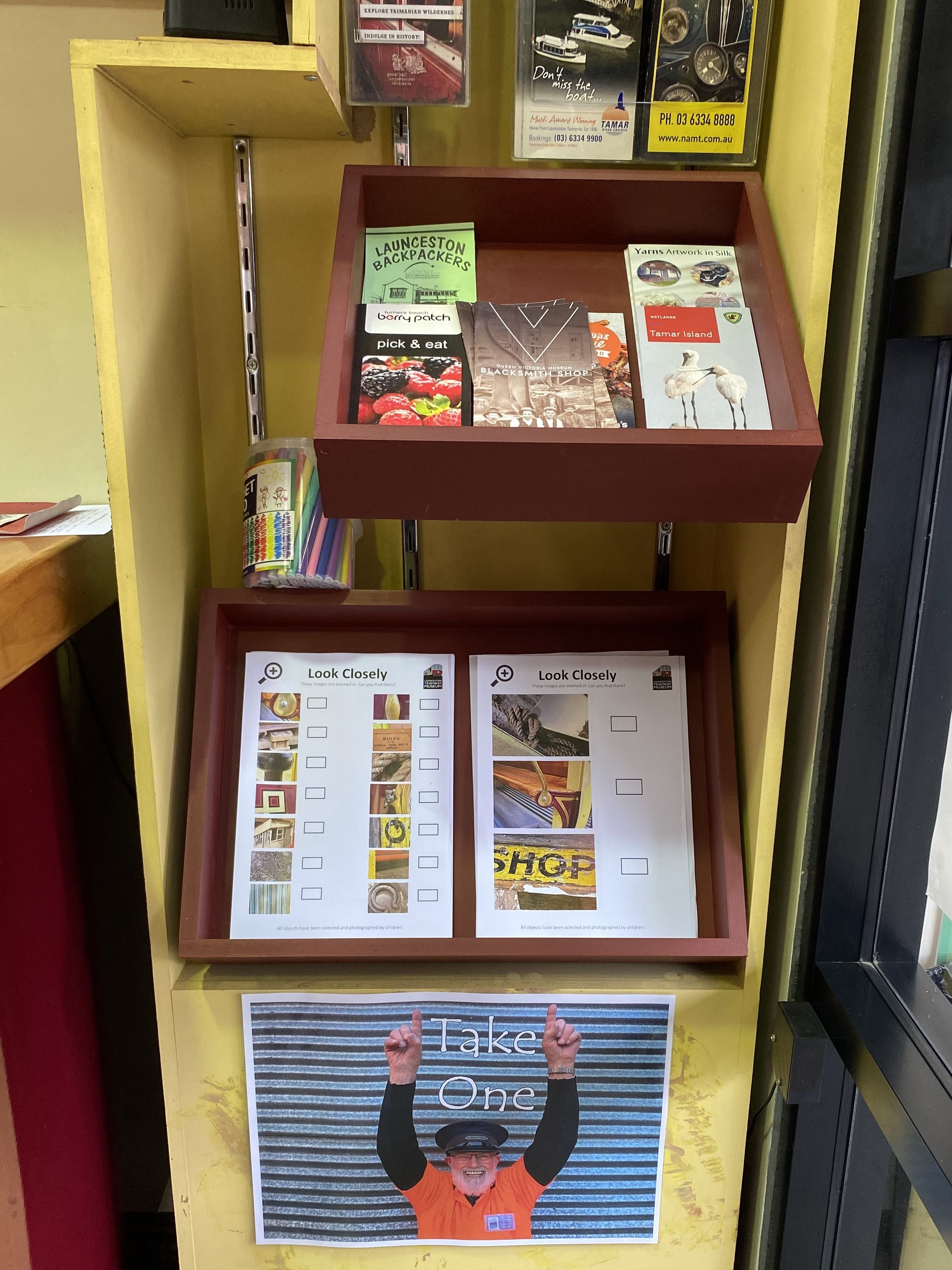 A yellow information stand at the Launceston Tramway Museum contains pamphlets and information sheets for various initiatives but prominent on the stand are the 'Look Closely' activity sheets designed by Limitless Learning participants. A photograph of the station master pointing towards the sheets saying 'Take One' is stuck to the bottom of the stand. 