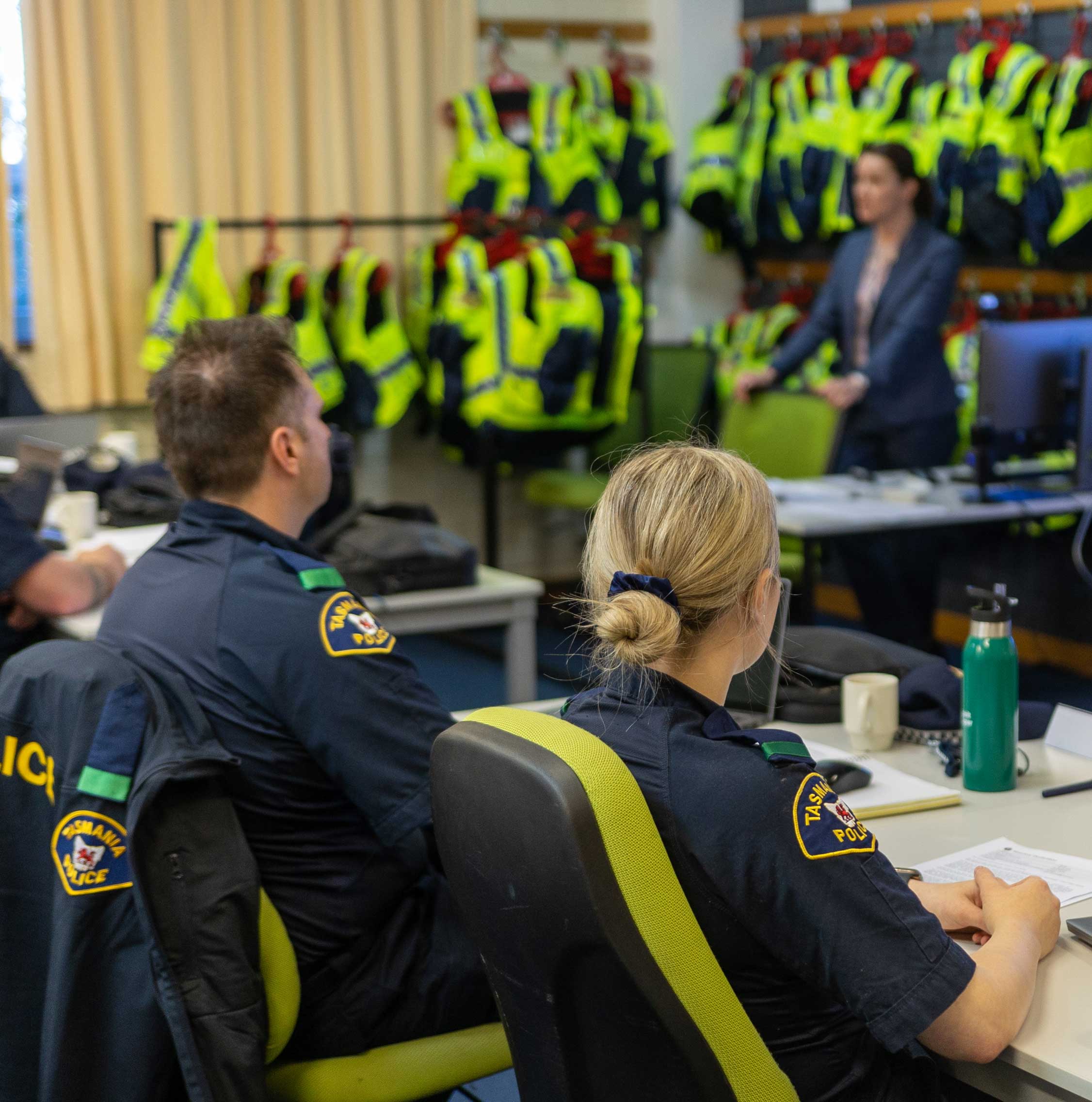 Group of police officers in a briefing room with high-visibility vests hanging in the background.