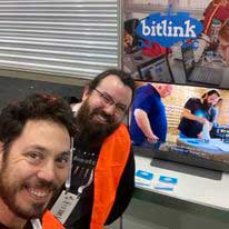 Two individuals in a room watching a demonstration on a television screen.