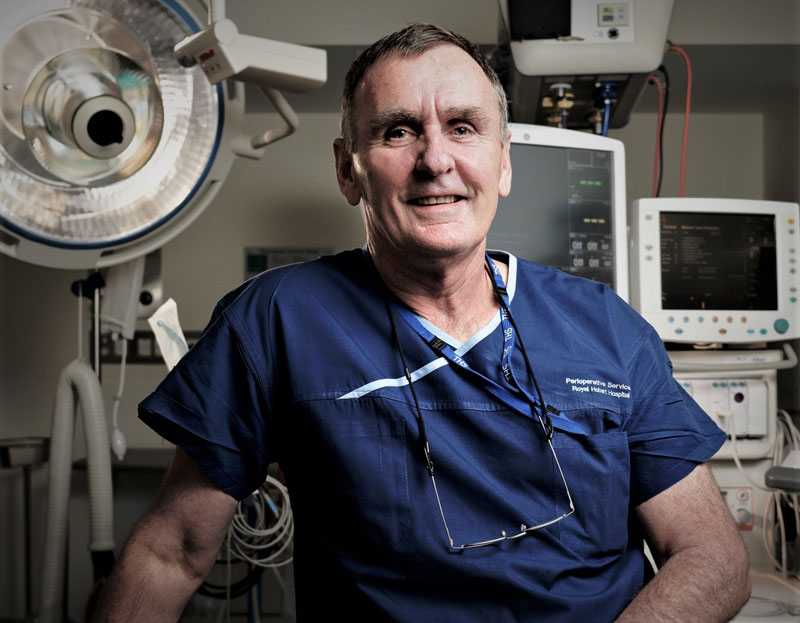 Marcus Skinner wearing dark blue medical scrubs with a stethoscope and glasses hanging from the neckline, seated in a hospital operating room surrounded by surgical lights and monitoring equipment.