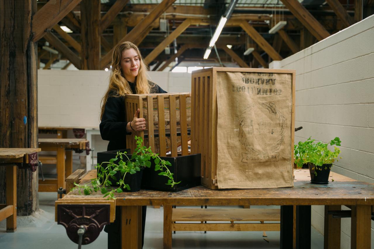A young woman dressed in all black, with fair skin and long dark-blonde hair, holds a planter overflowing with green herbs as she assembles a sharing-point for fresh produce, which resembles a wooden crate with a hessian sheet over the front.