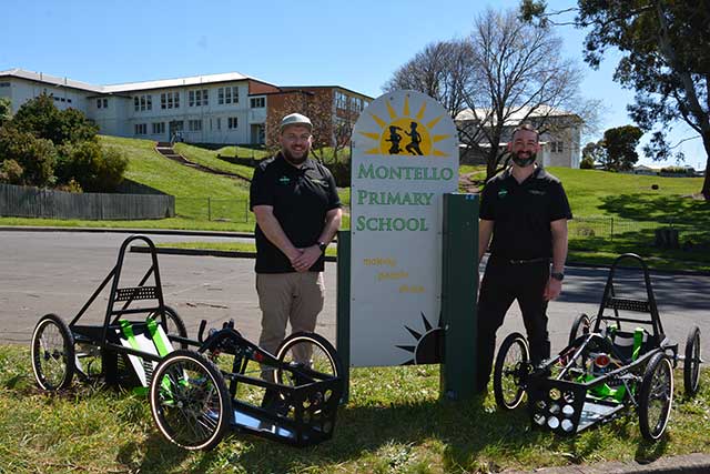 Two individuals standing beside a sign for Montello Primary School with three pedal-powered carts in the foreground.