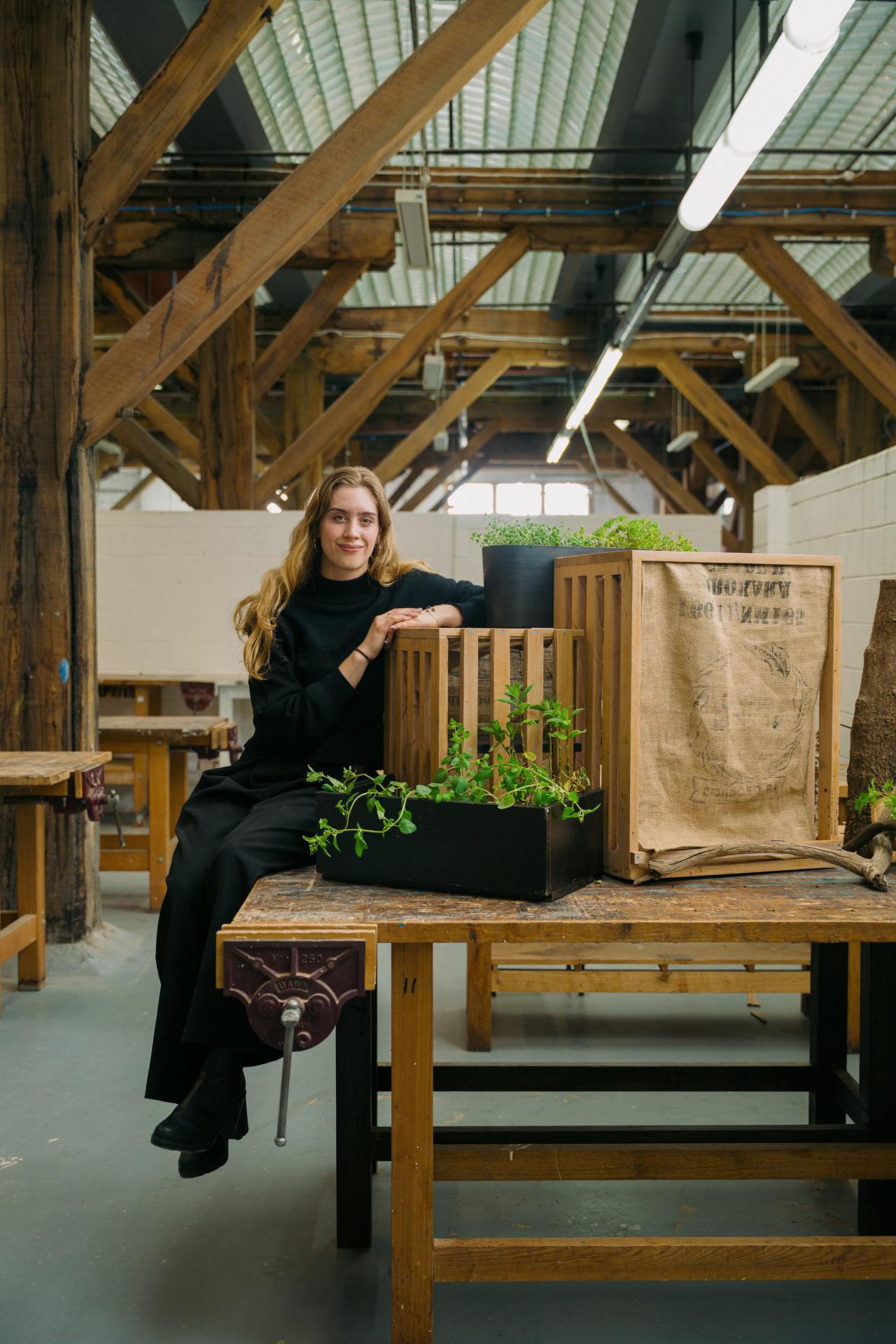 A young woman dressed in all black, with fair skin and long dark-blonde hair, sits on the edge of a table, which holds a planter overflowing with green herbs and a sharing-point for fresh produce, which resembles a wooden crate with a hessian sheet over the front.