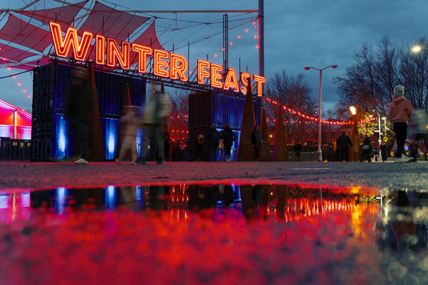 Winter Feast entrance at Dark Mofo Hobart, featuring glowing neon sign, crowds walking past food stalls, red festival lighting, and reflections on wet pavement at night.